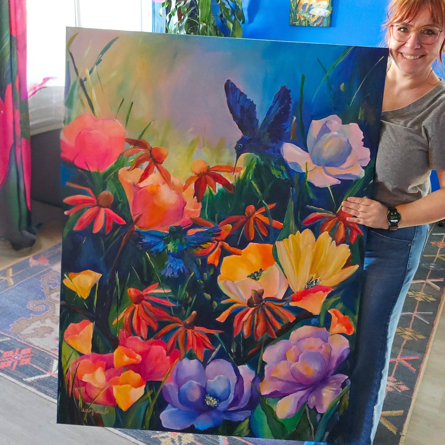 arge statement piece of hummingbirds in flight in a colorful floral garden perfect canvas wall art for a maximalsit living room or boutique hotel Artist Leah holding her 36x48 original floral and hummingbird painting ‘To Micrathene,’ showing bright botanical colors and detailed blue hummingbirds.